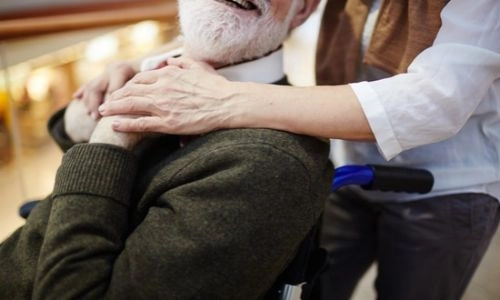 Une personne âgée en fauteuil roulant, souriante, tient la main posée sur son épaule par une proche qui se tient derrière elle.