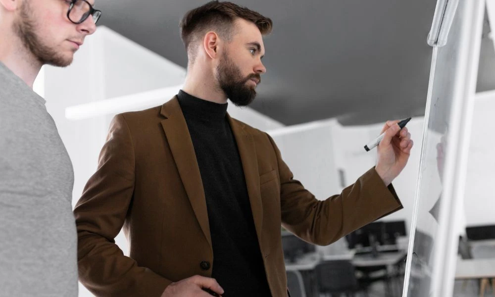 Un homme écrit sur un tableau blanc sous le regard attentif d’un collègue.