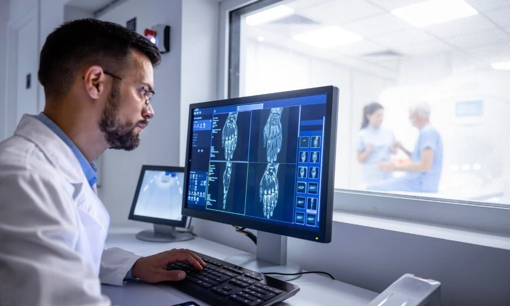 Un radiologue analyse des images d’IRM sur un écran, tandis qu’une soignante accompagne un patient dans la salle d’examen derrière la vitre.