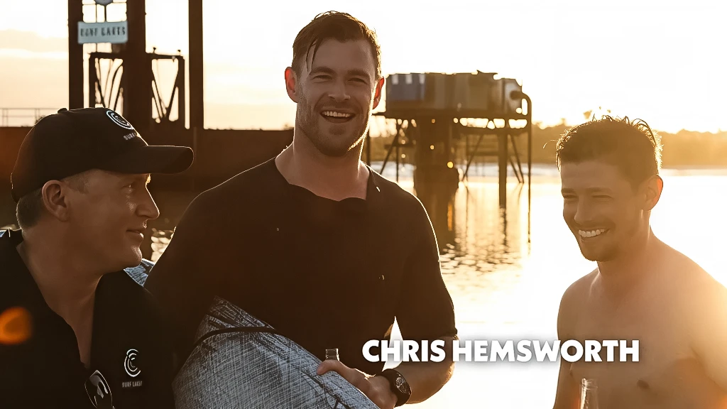Chris Hemsworth stands laughing between two men near water at sunset.