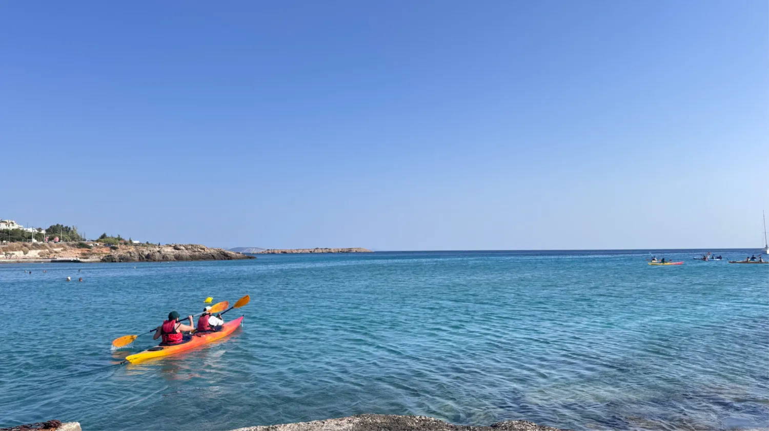 Two people paddling a kayak in clear turquoise water off a rocky coastline
