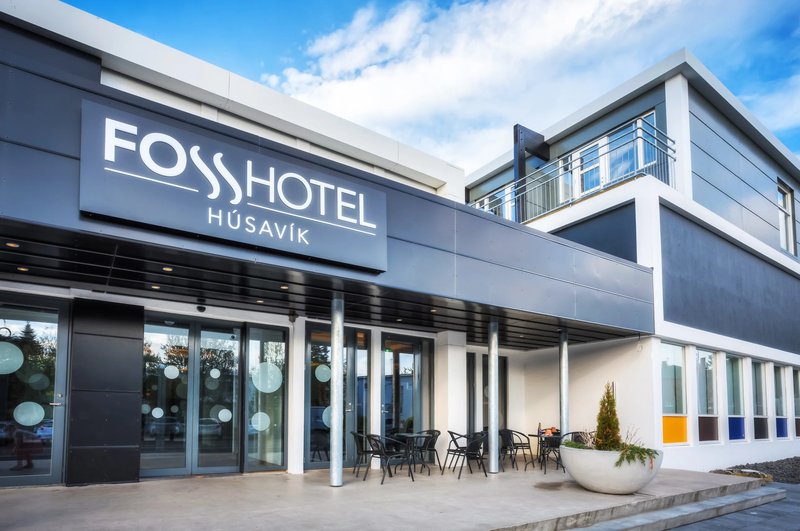 Entrance of Fosshótel Húsavík with the illuminated hotel sign above glass doors, a small outdoor seating area and a planter under a blue sky