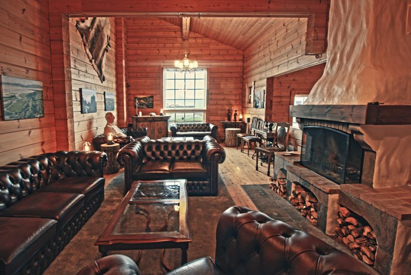 Wood-paneled lounge at Hótel Breiðdalsvík with brown leather Chesterfield sofas, a glass coffee table and a large stone fireplace with stacked logs