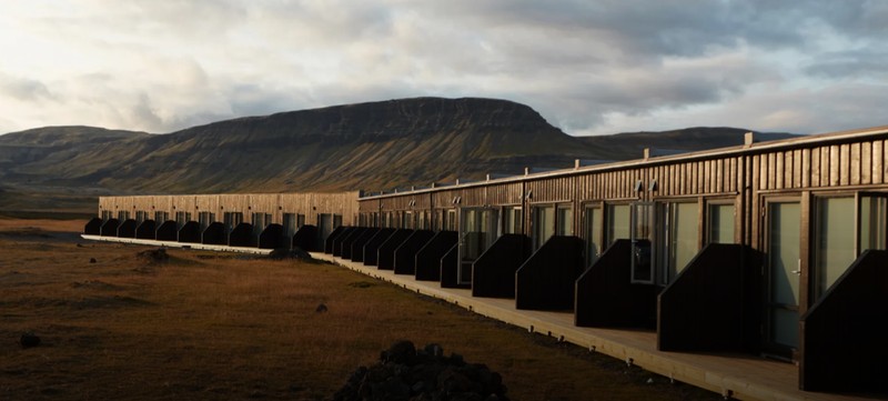 Long single-storey row of dark timber-clad rooms at Fosshótel Núpar, each with a glass door and wooden deck, in front of a flat-topped mountain in evening light