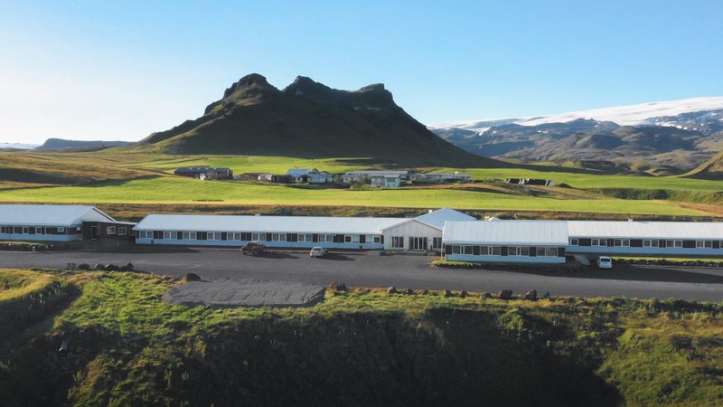 Hótel Dyrhólaey, long low white-roofed buildings on green farmland below a craggy mountain, with a snow-capped glacier in the distance