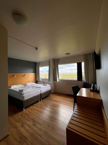 Guest room at Hótel Jökull with two single beds pushed together, an oak headboard, wood-effect floor and wide windows looking out over green fields and mountains
