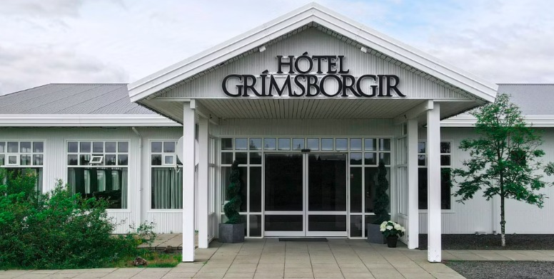 Entrance of Hótel Grímsborgir, a white single-storey building with a gabled porch, the hotel name above glass doors and a small tree and planters in front