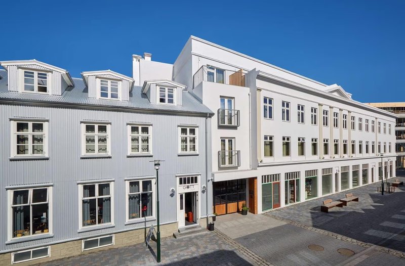 Exterior of Konsúlat Hotel Reykjavík, a white modern building next to a historic corrugated-iron house on a cobbled pedestrian street under a clear blue sky