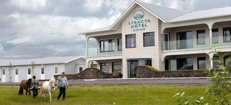 Exterior of Stracta Hotel Hella, a two-story cream building with balconies and a stone wall, with two people leading Icelandic horses across the grass in front