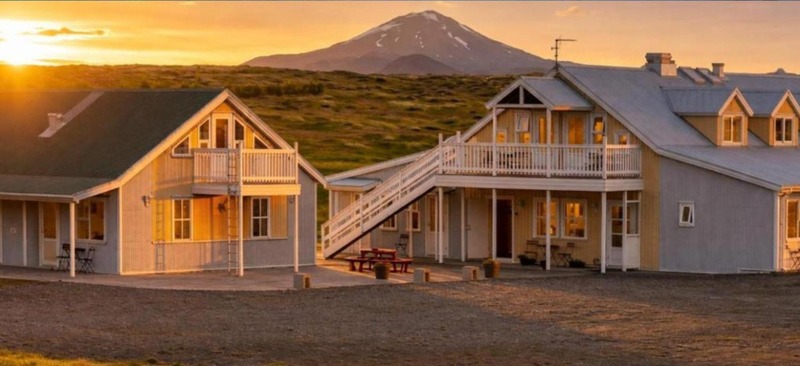 Hotel Lækur buildings with wooden balconies and an outdoor staircase at sunset, with snow-capped Hekla volcano rising behind