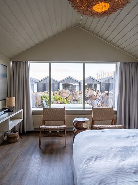 Guest room at The Hill Hotel Flúðir with a vaulted white ceiling, two woven chairs and a large window looking out at lava rock and dark cabins