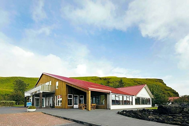 Hotel Katla in Vik with a timber-clad main building, red metal roof and restaurant wing, set against a green hillside and a blue sky with clouds