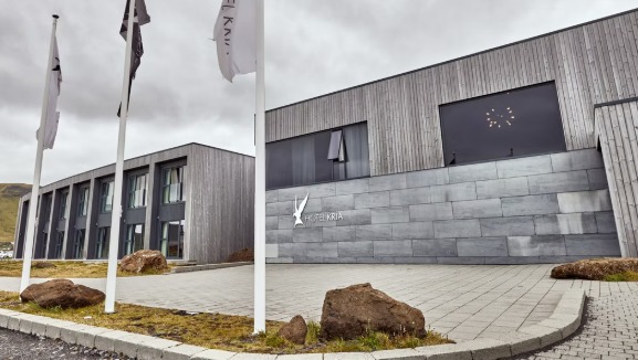 Entrance of Hotel Kría in Vík with a gray stone facade and timber-clad wings, flagpoles and lava boulders in front under an overcast sky