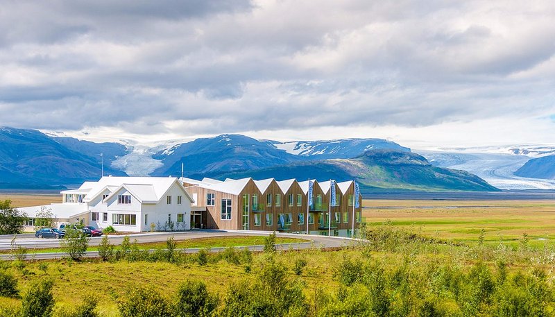 Fosshotel Vatnajokull with a white main building and a timber-clad wing with pitched gables, set on a plain beneath mountains and glacier tongues of Vatnajokull