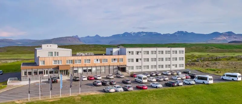 Aerial view of Fosshotel Stykkisholmur, a white and timber-clad hotel with a large parking lot, green fields and a mountain range behind