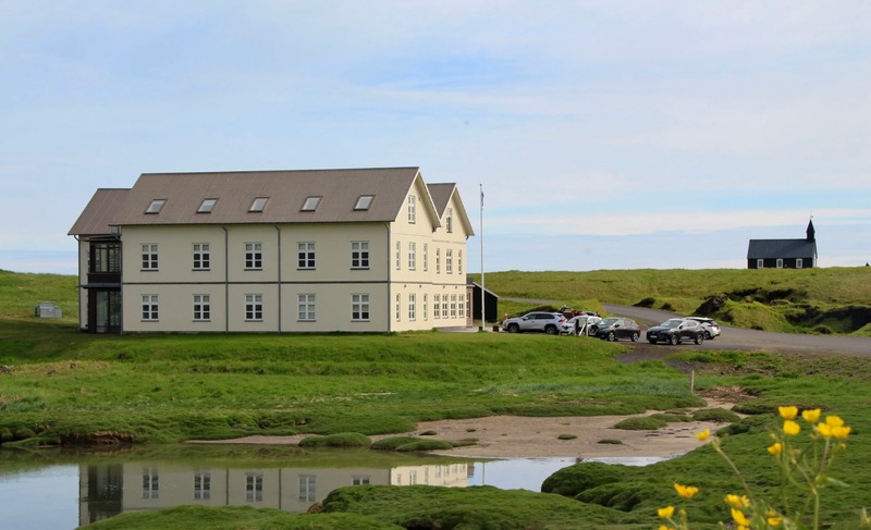 Hotel Búðir, a cream two-story building with a brown roof beside a tidal pool and green lava moss, with the black Búðir church on the hill behind