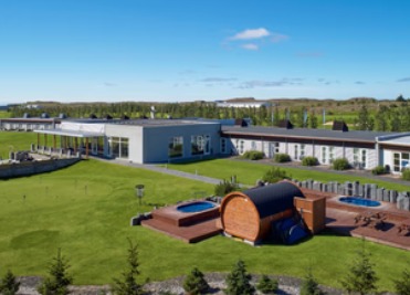 Aerial view of Hótel Hamar, low gray hotel wings on a green lawn with two hot tubs and a barrel sauna on a wooden deck in the foreground
