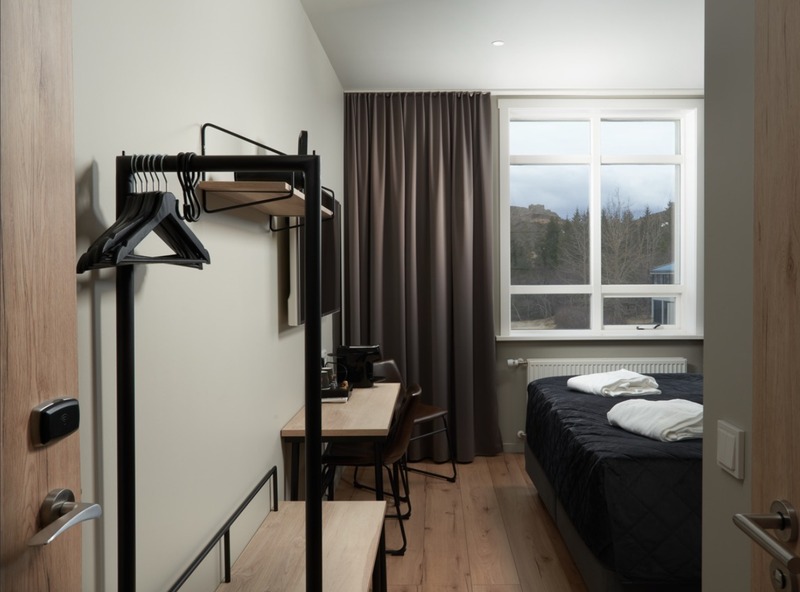 Guest room at Hótel Varmaland with a black-framed clothing rack, wooden desk, dark double bed with white towels, and a window looking out to trees and a hillside