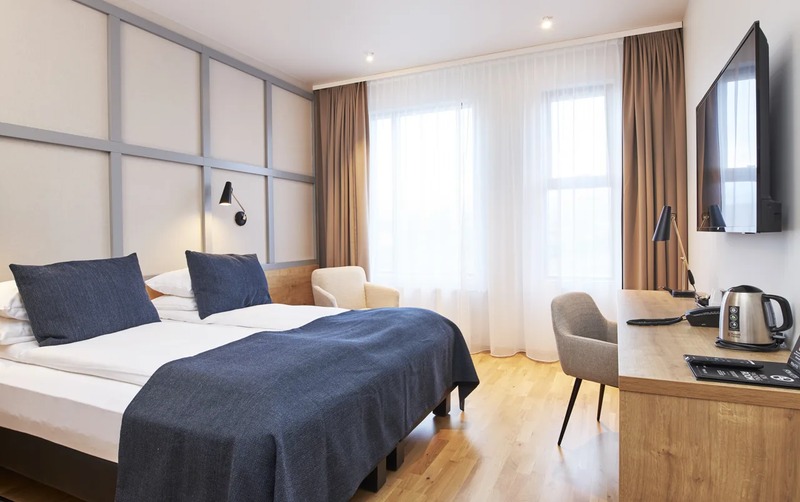 Double room at Fosshótel Reykholt with a navy blanket and pillows, gray paneled wall, tall sheer-curtained windows, a wooden desk with kettle, and light oak flooring