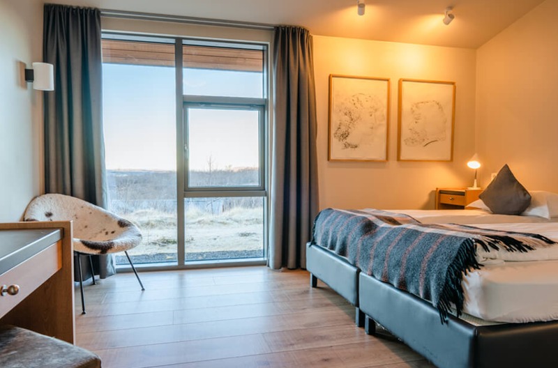 Guest room at Hótel Húsafell with twin beds under a wool throw, a sheepskin chair, and a floor-to-ceiling window overlooking open moorland at sunset