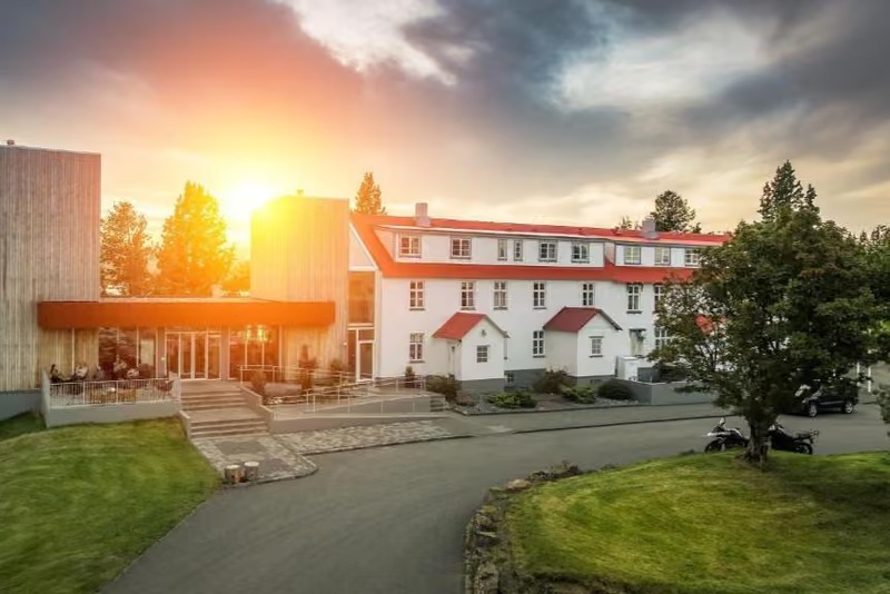Exterior of Lake Hotel Egilsstaðir, a white building with a red roof and modern wood-clad wing, lit by a golden sunset behind autumn trees