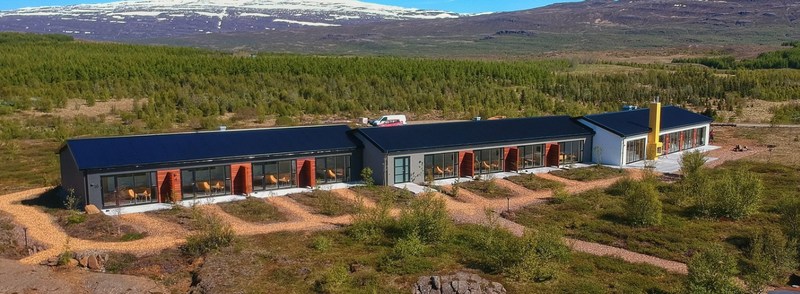 Aerial view of Hotel 1001 Nótt, a long low building with dark roofs, floor-to-ceiling windows and a yellow chimney, set in moss and birch forest with snowy mountains behind