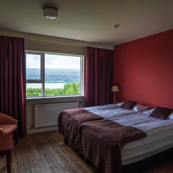 Guest room at Hotel Hallormsstaður with twin beds, burgundy curtains and a red accent wall, and a window looking over green trees to the lake and hills