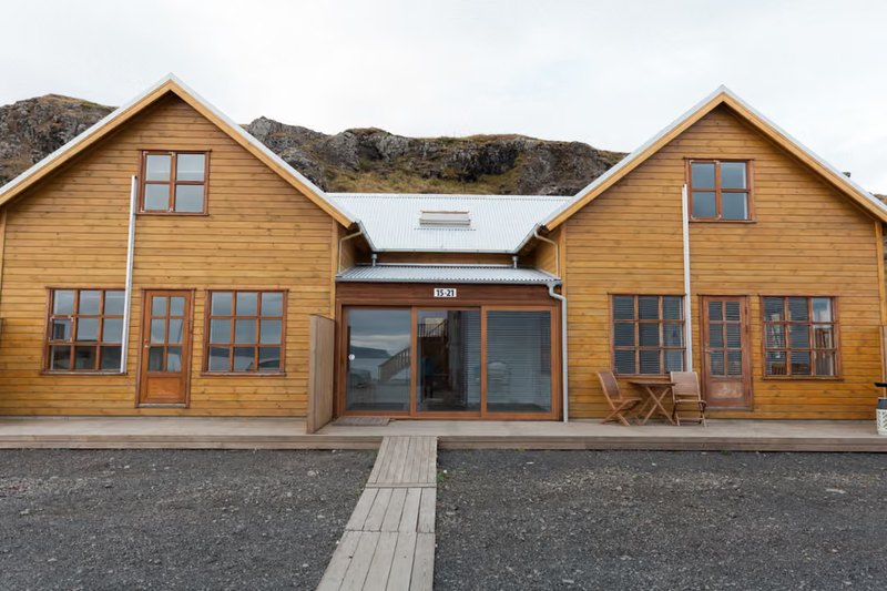 Front of Malarhorn guesthouse, two gabled golden-wood buildings with a glass entrance between them and a boardwalk across dark gravel, with a rocky hillside behind