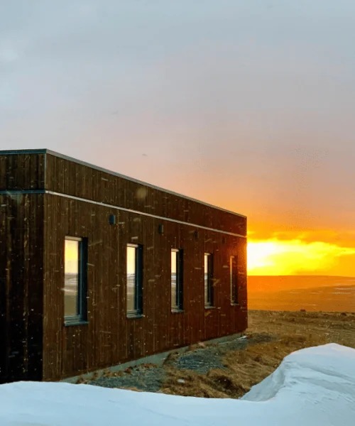 Exterior of Hótel Laxá, a dark timber-clad building with tall narrow windows, with a glowing orange sunset over open land and a snowdrift in the foreground