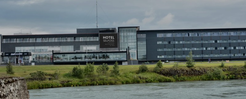 Exterior of Hotel Selfoss Restaurant and Spa, a long dark gray multi-story building with glass sections, seen across the river and grassy bank under a cloudy sky