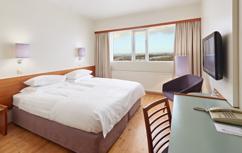 Double room at Hérað Berjaya Hotel with a white-linen bed, wood headboard panel, wall sconces, a purple armchair, a desk, and a wide window with a view over the town