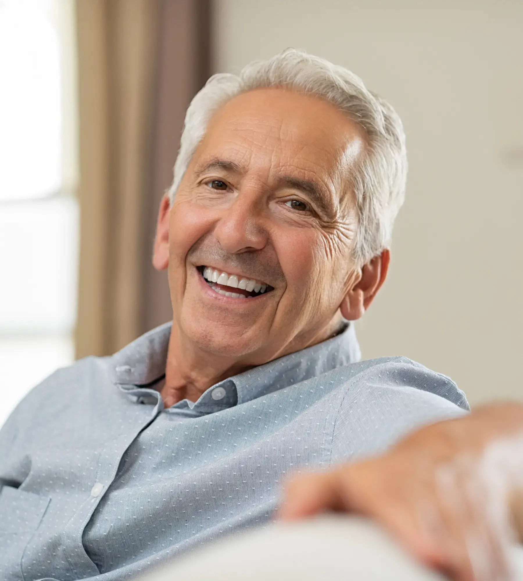 Older man smiling while sitting indoors, looking directly at the camera.