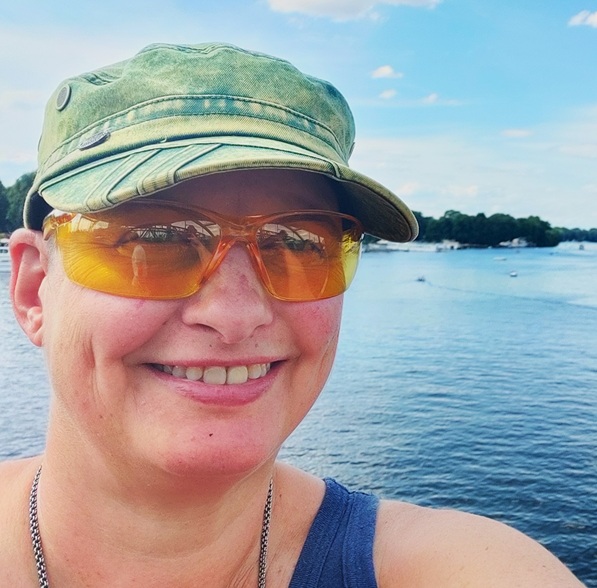 Portrait of Dr. GAl Schkolnik, Aut2Aut founder and CEO, a smiling white nonbinary person in a blue tank top, green cap and yellow protective glasses with a river and trees in the background