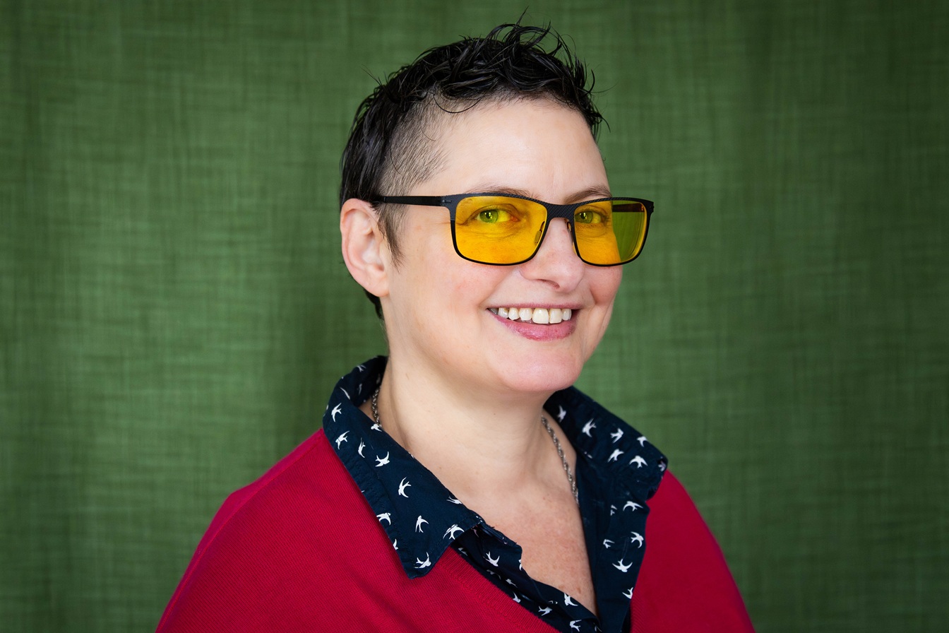 Portrait of Dr. GAl Schkolnik, Aut2Aut founder and CEO, a smiling white nonbinary person in a blue tank top, green cap and yellow protective glasses with a river and trees in the background