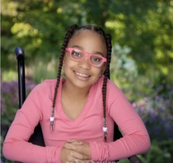 Young bald girl outside wearing pink dress smiling while touching tree branch with a river in the background.