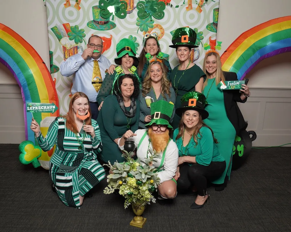 Family wearing green standing in front of a wall with illustrations of beer, shamrocks, and horseshoes holding paper cutouts of various St Patricks Day themed objects.