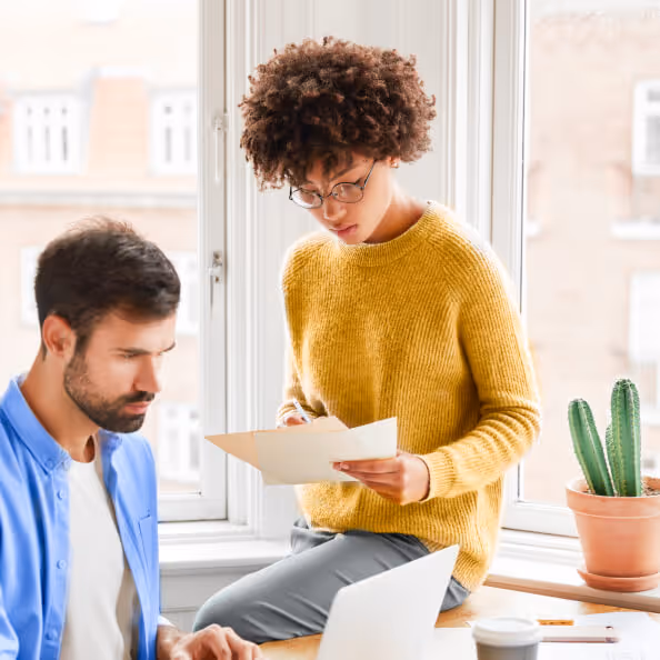 Woman sitting on desktop reading a paper document to her male colleague