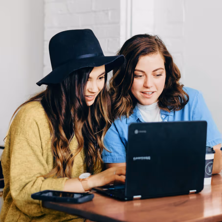 two women one wearing a black hat viewing a laptop