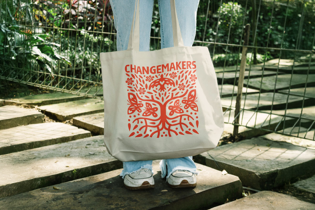 Canvas tote bag with red 'CHANGEMAKERS' text and decorative tree design held by person wearing light blue jeans and white sneakers outdoors.