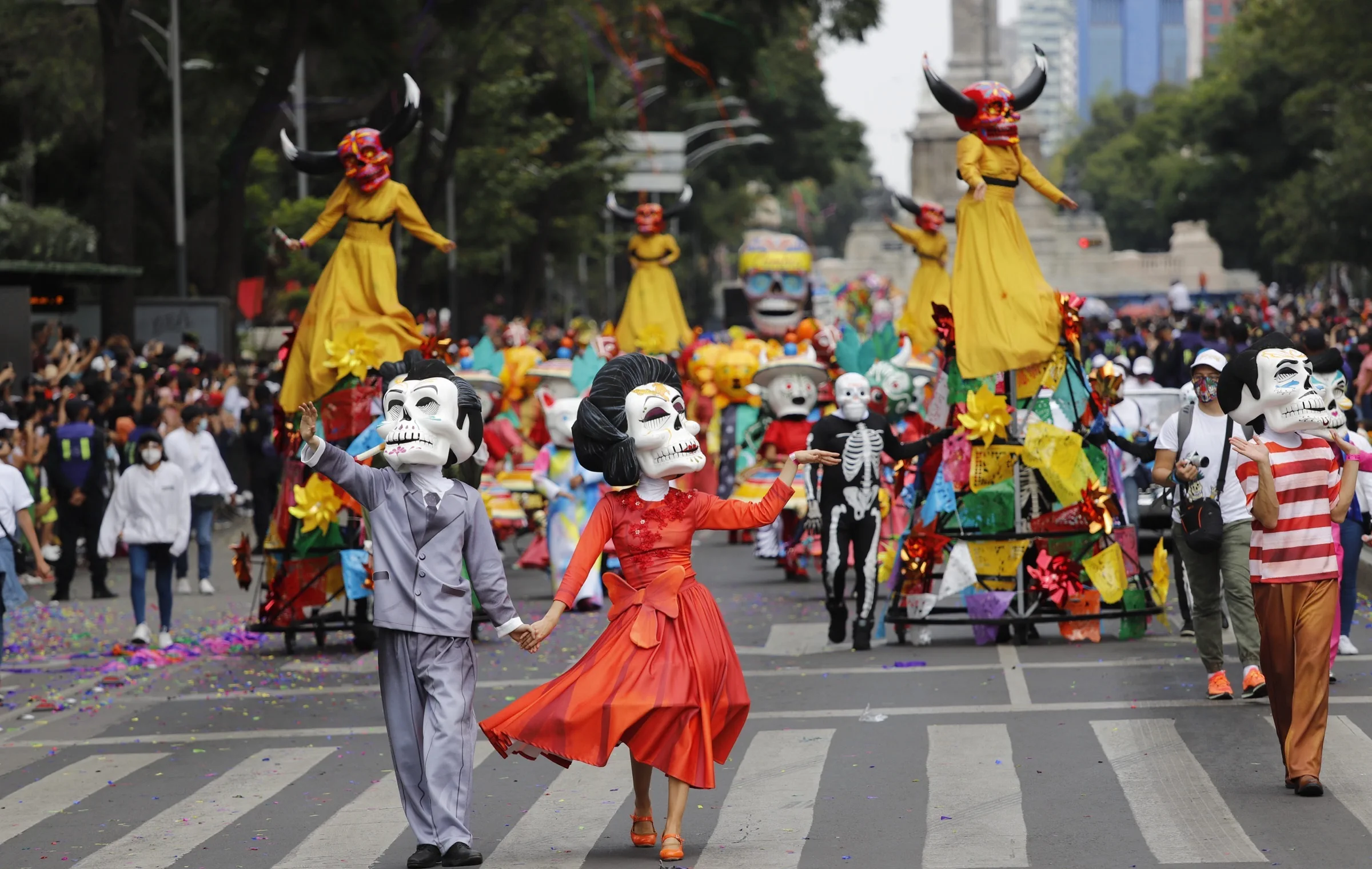 Day of the Dead parade featuring two people in the foreground wearing elegant outfits and skull masks, dancing in the street as large floats with Catrina figures and bull skull papier-mâché decorations move in the background, surrounded by spectators.