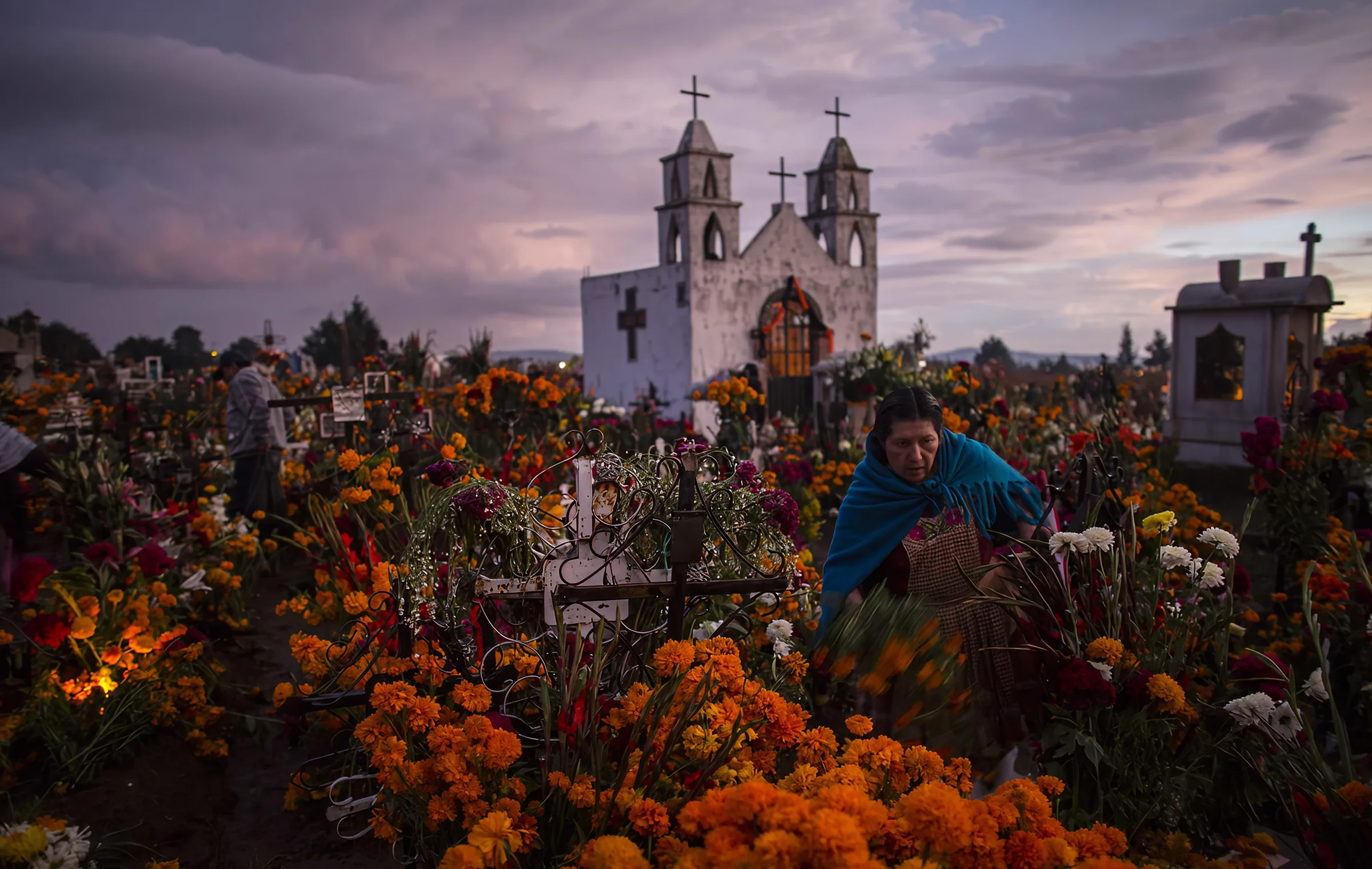 A woman in a blue shawl decorates a grave with cempasúchil flowers in a cemetery at dusk, with a white church visible in the background under a dramatic sky.