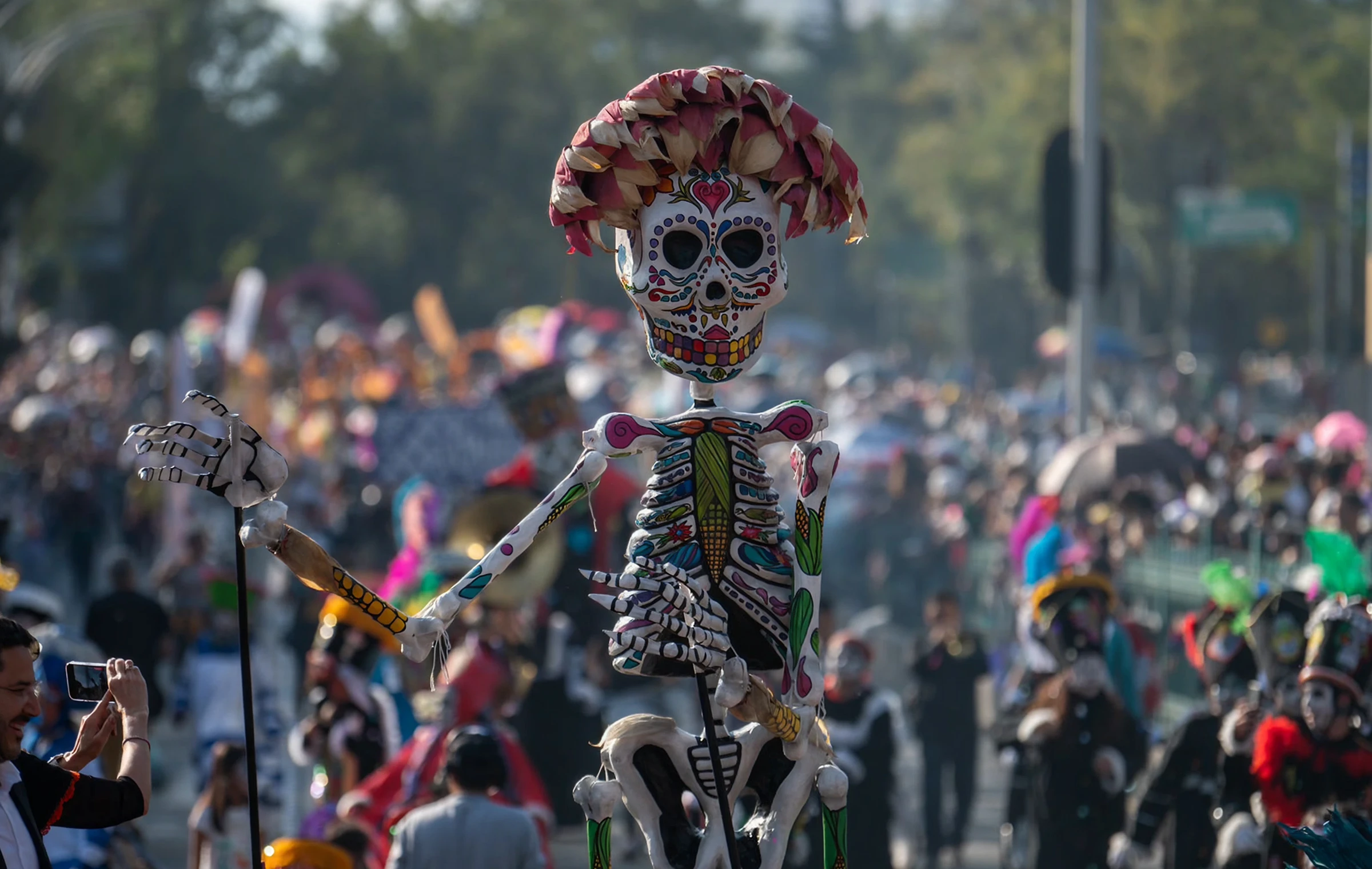 Close-up of a large skeleton puppet, colorfully decorated in a sugar skull style with a paper flower headdress, during a Day of the Dead parade with a blurred crowd in the background.