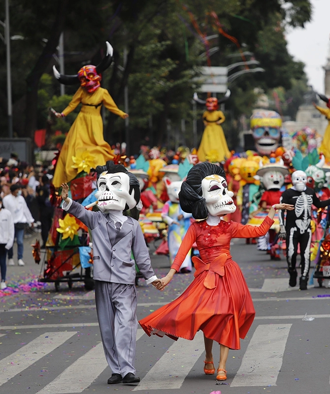 Una pareja disfrazada con máscaras de calavera, él de traje gris y ella de vestido rojo, bailan tomados de la mano en un desfile del Día de Muertos, rodeados de carrozas y otros participantes.