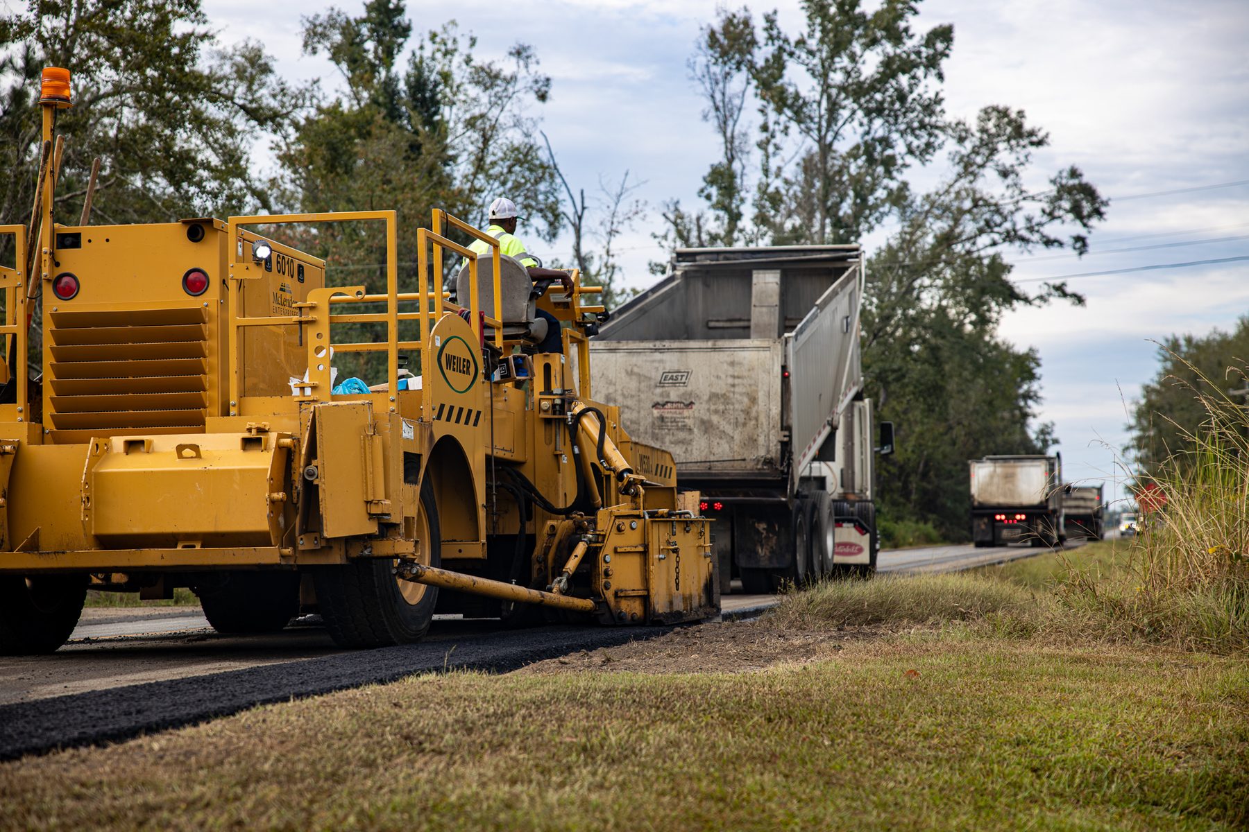 **Alt Text:** A road construction crew is paving asphalt along Cedar Crossing Road in Vidalia, GA. Heavy machinery, including a yellow paver, is actively involved in the widening project, with dump trucks lined up for material delivery. ...