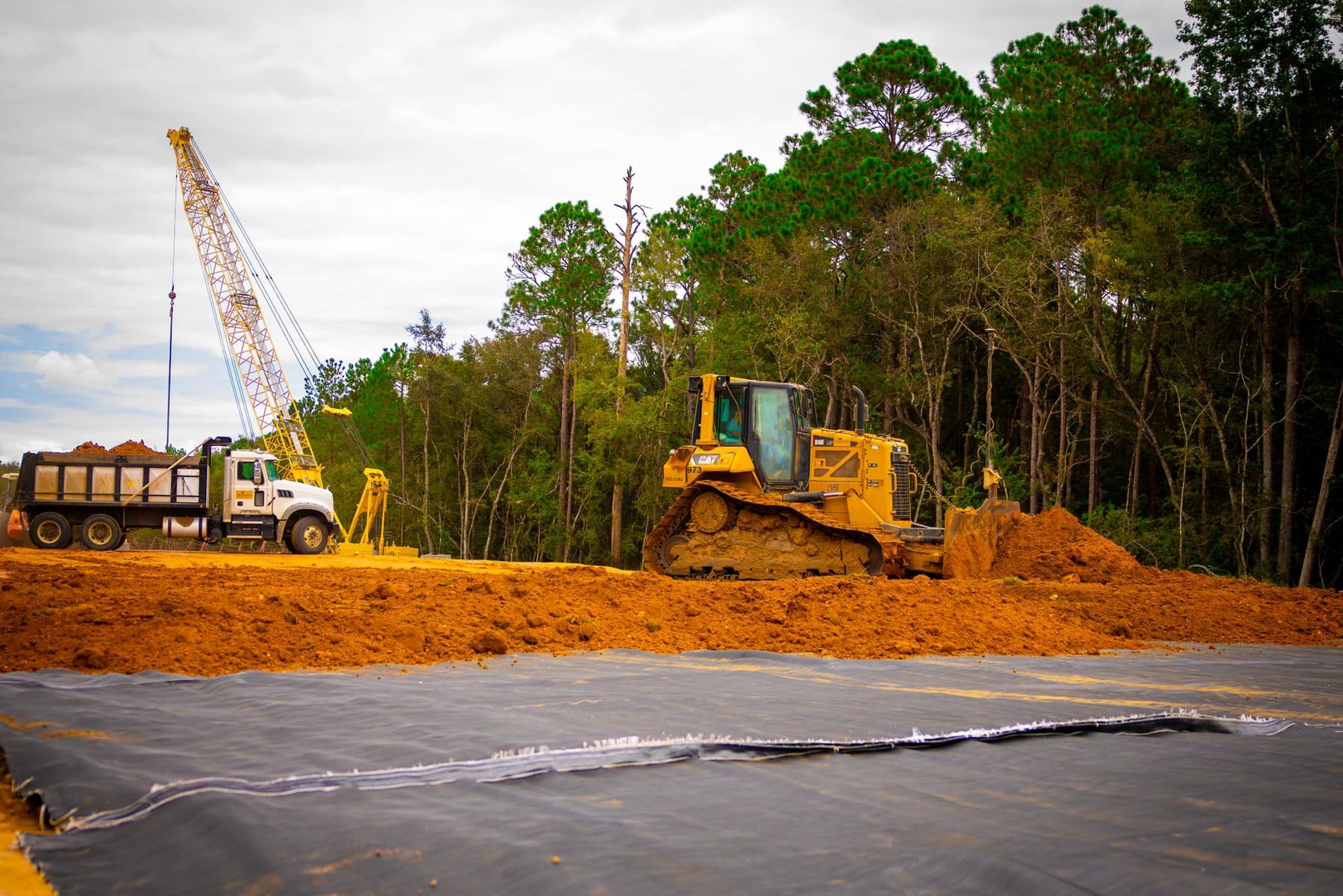 **Alt Text:** A Caterpillar bulldozer is actively moving earth on the construction site of the US 1 Bypass in Toombs County, GA. Behind it, a crane lifts materials while a dump truck is parked nearby, showcasing our commitment to geotext...