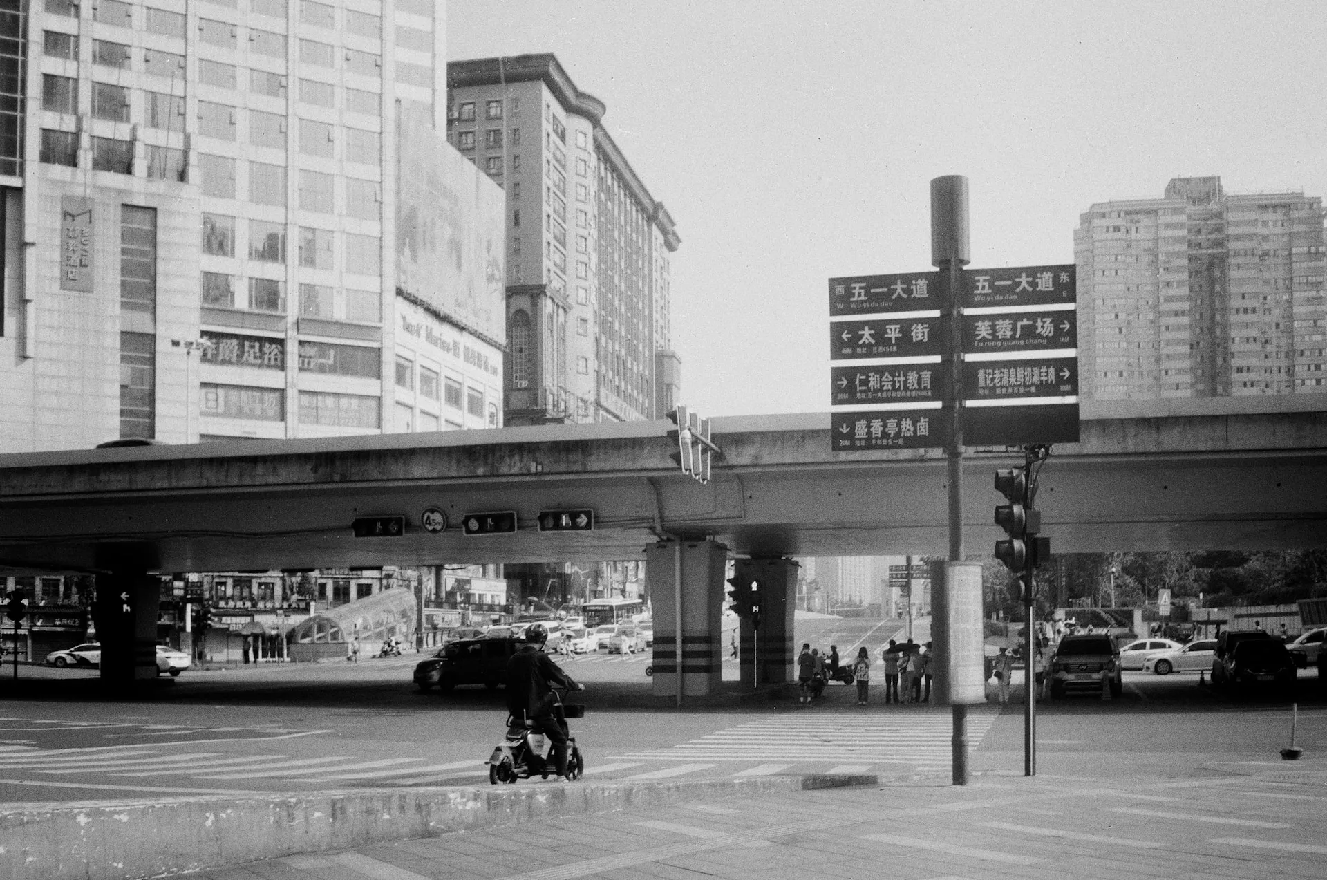 A black and white photograph of a person riding a scooter in an urban area