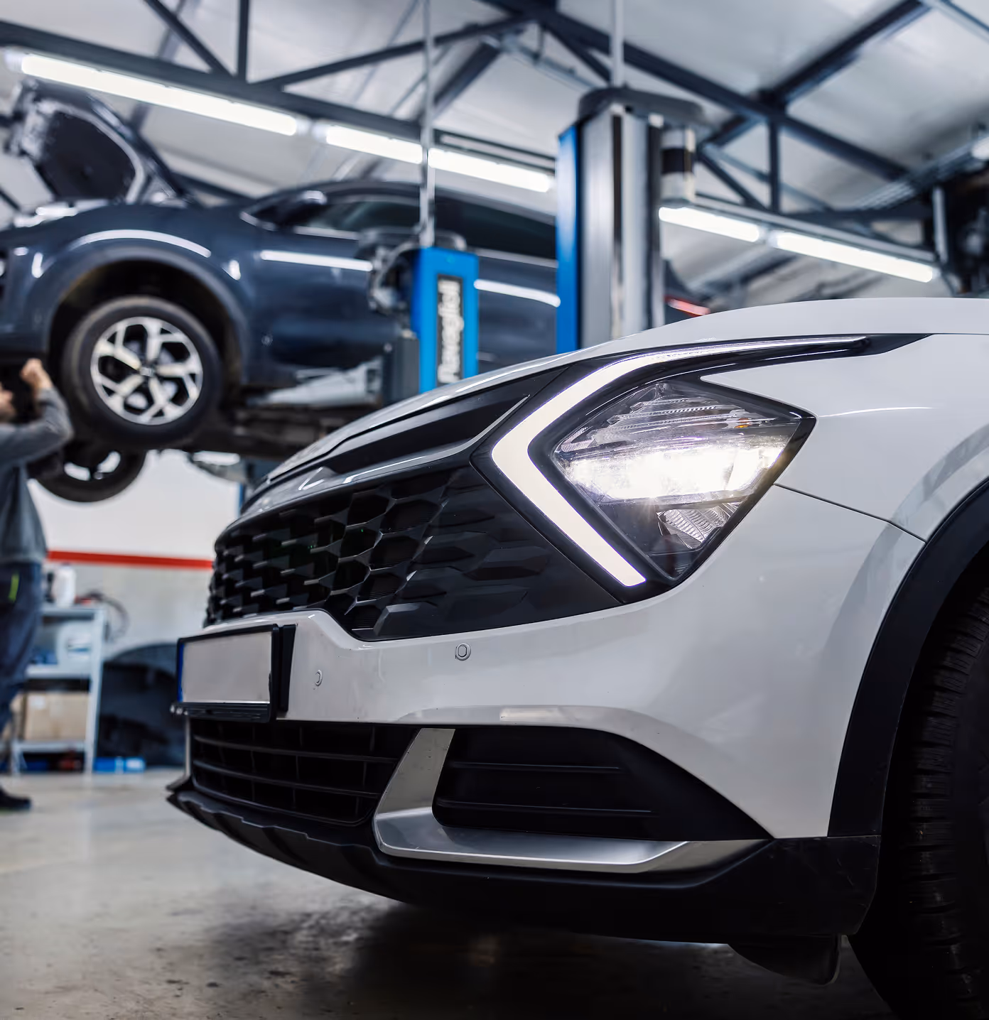 The front end of a recently repaired vehicle in a brightly lit autobody shop. In the background another vehicle is on a hoist being repaired by a tech.