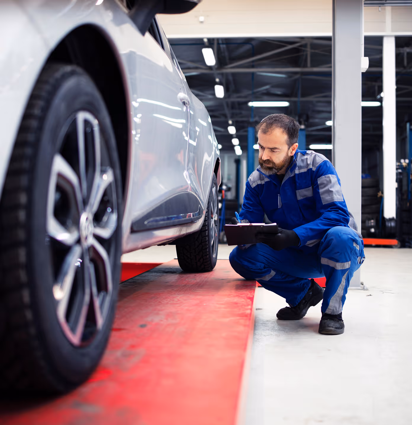 Repair technician crouching next to a car that’s been repaired, he’s holding a clipboard and inspecting the vehicle near the seam of the doors.