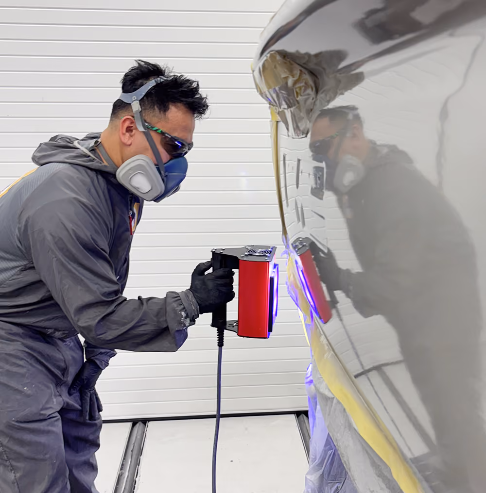 A technician in a gray jumpsuit, respirator mask, and safety glasses uses a handheld red UV curing lamp on the side of a vehicle in a body shop.