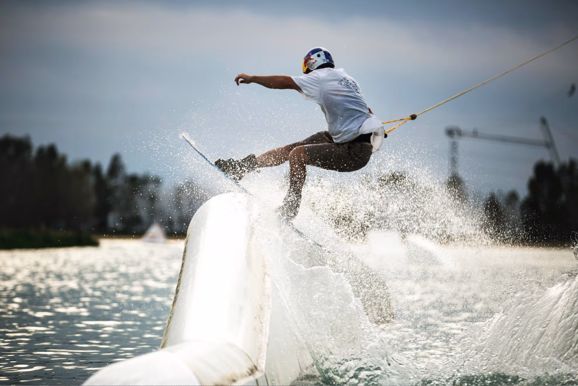Person wakeboarding on water ramp, creating a large splash while wearing a helmet and white t-shirt.